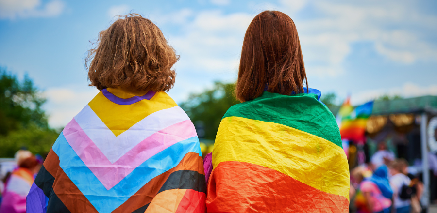 Two people with their backs to the camera, wearing LGBTQIASB+ flags over their shoulders